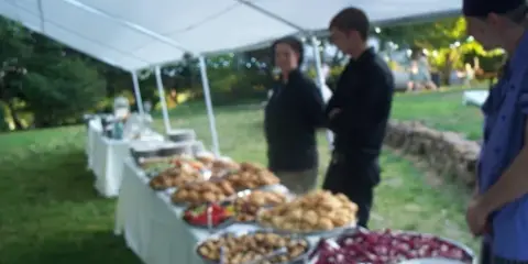 Outdoor catering event with a long table of food and staff under a white tent.