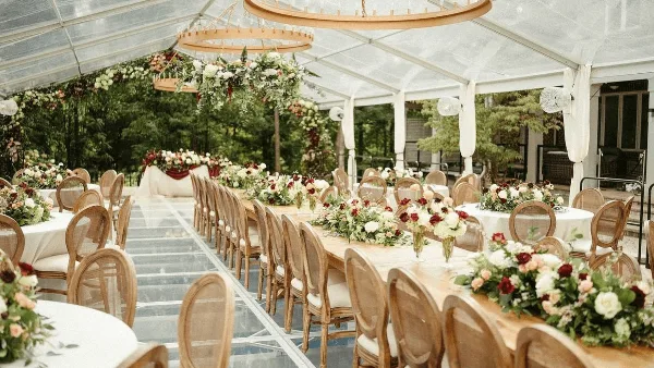 A wedding reception venue featuring long tables with floral centerpieces, wooden chairs, and hanging chandeliers under a clear tent.