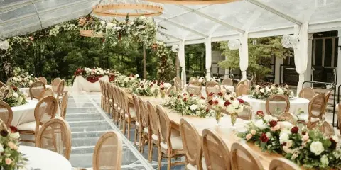 A wedding reception venue featuring long tables with floral centerpieces, wooden chairs, and hanging chandeliers under a clear tent.