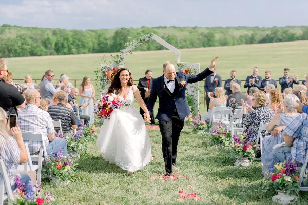 A bride and groom walking down the aisle during an outdoor wedding ceremony in a grassy field with guests seated on either side.