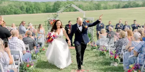 A bride and groom walking down the aisle during an outdoor wedding ceremony in a grassy field with guests seated on either side.
