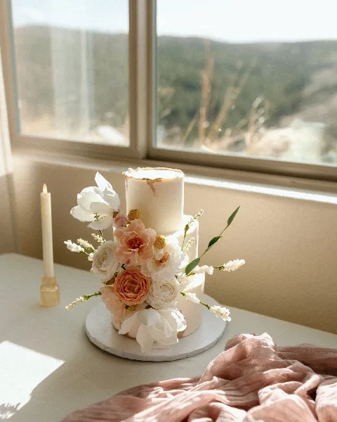 A three-tier white wedding cake decorated with pink and white flowers and gold accents, sitting on a table near a window.