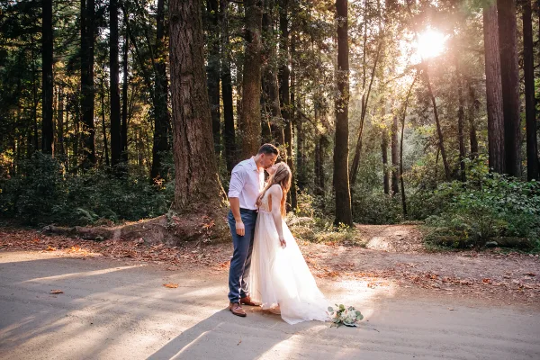 A bride and groom sharing a kiss on a forest path with sunlight filtering through the trees.