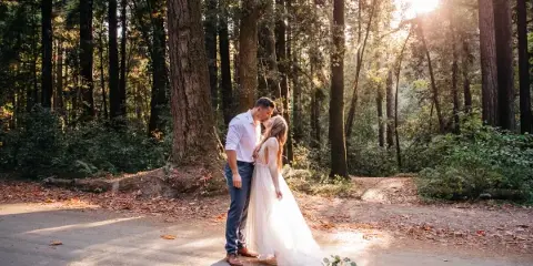 A bride and groom sharing a kiss on a forest path with sunlight filtering through the trees.