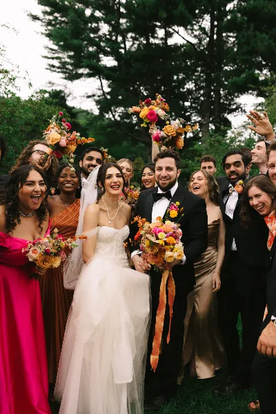 A bride and groom surrounded by their wedding party, holding colorful bouquets and smiling outdoors.