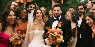 A bride and groom surrounded by their wedding party, holding colorful bouquets and smiling outdoors.