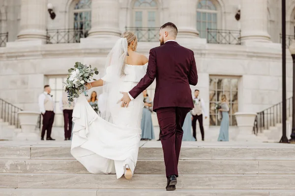 A bride and groom walking down stone steps in front of a classical building, with the bride holding a bouquet and the groom in a maroon suit.