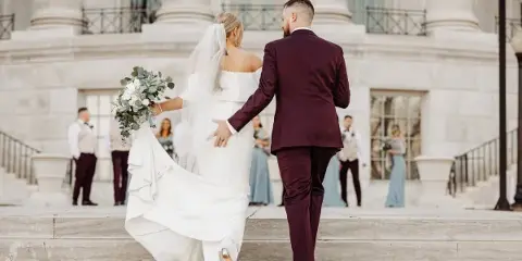 A bride and groom walking down stone steps in front of a classical building, with the bride holding a bouquet and the groom in a maroon suit.