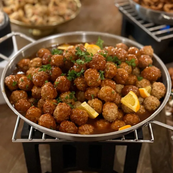 A large metal chafing dish filled with glazed meatballs garnished with parsley and lemon wedges, served at a catering event.