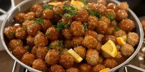 A large metal chafing dish filled with glazed meatballs garnished with parsley and lemon wedges, served at a catering event.