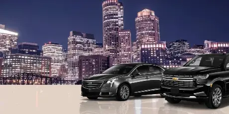 A black luxury sedan and a black SUV parked in front of the Boston city skyline at night.