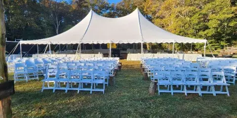 Outdoor wedding setup featuring a large white tent, rows of white folding chairs, and tables on a grassy lawn.