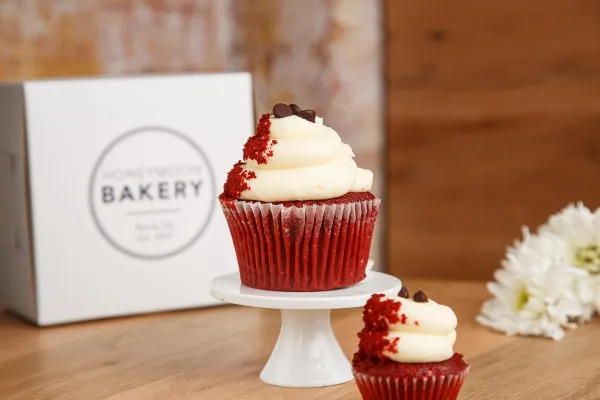 A red velvet cupcake with white frosting and chocolate chips on a white cake stand, with a Honeymoon Bakery box in the background.