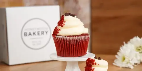 A red velvet cupcake with white frosting and chocolate chips on a white cake stand, with a Honeymoon Bakery box in the background.