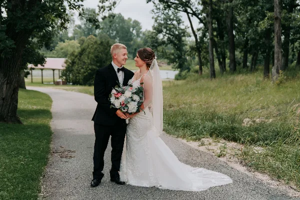A bride and groom standing on a gravel path outdoors, with the bride holding a bouquet of white and pink flowers.