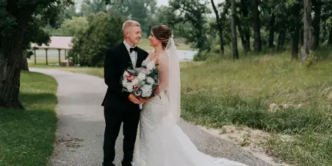 A bride and groom standing on a gravel path outdoors, with the bride holding a bouquet of white and pink flowers.