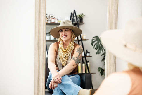 A smiling woman with a tattooed arm and a hat sits in a salon chair, with a mirror and shelves of hair products visible in the background.