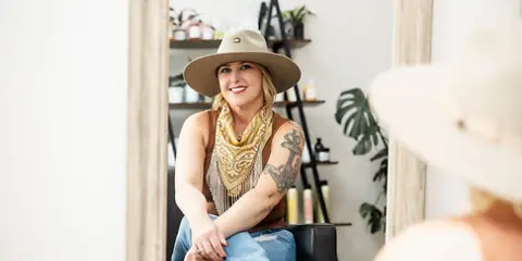 A smiling woman with a tattooed arm and a hat sits in a salon chair, with a mirror and shelves of hair products visible in the background.
