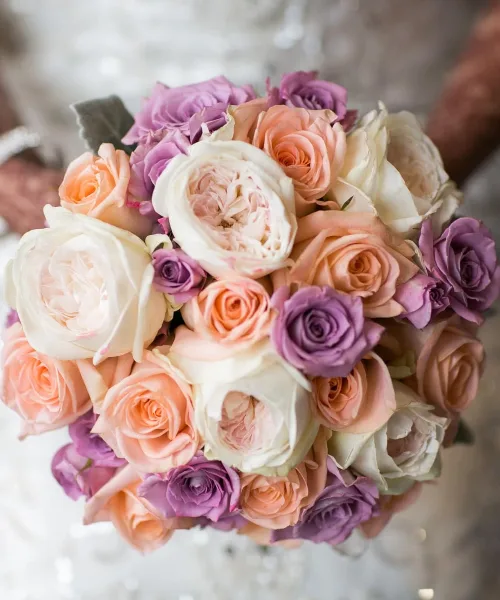 A close-up photograph of a round bridal bouquet composed of peach, cream, and purple roses and garden roses.