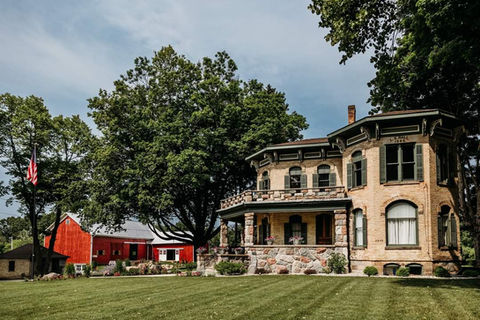 A historic stone house with a porch and a red barn situated on a grassy lawn.