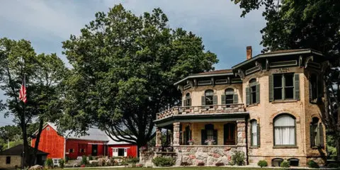 A historic stone house with a porch and a red barn situated on a grassy lawn.