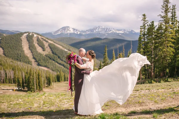A bride and groom kissing in a mountainous outdoor setting with a flowing white dress and bouquet.
