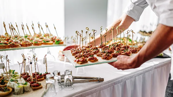 A caterer in a white shirt places a tray of skewered appetizers on a white-draped event table filled with other canapés and glassware.