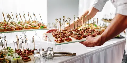 A caterer in a white shirt places a tray of skewered appetizers on a white-draped event table filled with other canapés and glassware.