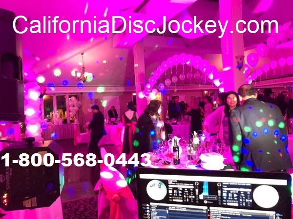 A DJ booth with a Pioneer controller and speakers set up in a wedding reception venue with guests dancing under pink and blue lights.