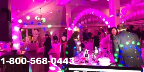 A DJ booth with a Pioneer controller and speakers set up in a wedding reception venue with guests dancing under pink and blue lights.