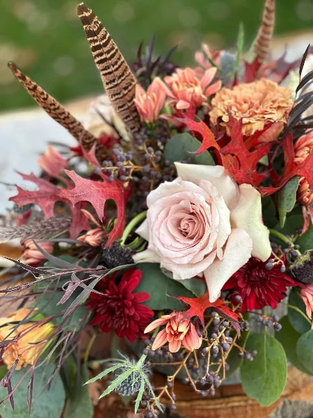 A rustic floral arrangement featuring a central cream rose, red maple leaves, and brown feathers.