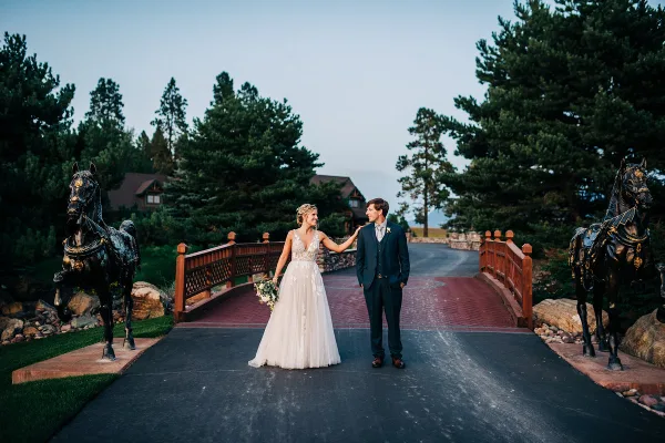 A bride and groom standing on a paved path flanked by two large bronze horse statues, with a wooden bridge and pine trees in the background.