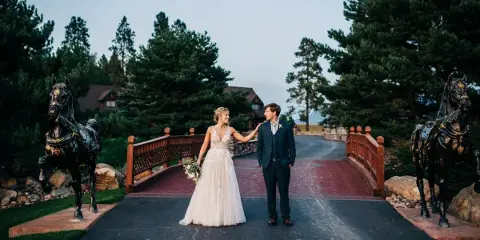 A bride and groom standing on a paved path flanked by two large bronze horse statues, with a wooden bridge and pine trees in the background.
