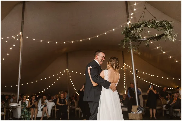A bride and groom dancing under string lights and a floral chandelier inside a large event tent during a wedding reception.