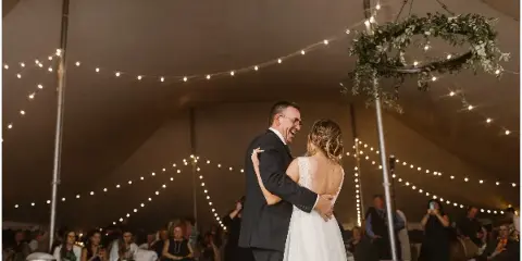 A bride and groom dancing under string lights and a floral chandelier inside a large event tent during a wedding reception.