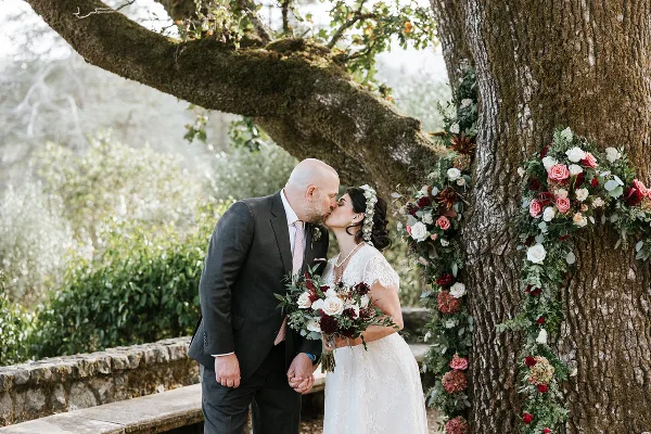 A bride and groom sharing a kiss next to a large tree decorated with floral garlands and a stone wall in the background.