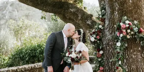 A bride and groom sharing a kiss next to a large tree decorated with floral garlands and a stone wall in the background.