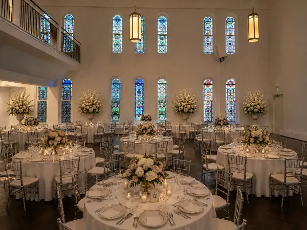 Interior view of a wedding reception hall with round tables, white linens, floral centerpieces, and tall stained glass windows.