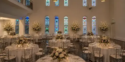 Interior view of a wedding reception hall with round tables, white linens, floral centerpieces, and tall stained glass windows.