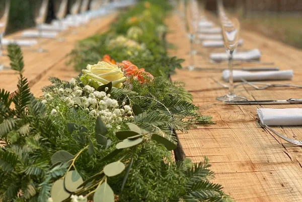 A long wooden dining table set with white napkins, silverware, and a lush green floral centerpiece.