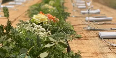 A long wooden dining table set with white napkins, silverware, and a lush green floral centerpiece.