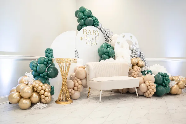 A professionally styled event backdrop featuring a green, gold, and silver balloon arch, a white velvet loveseat, and a gold pedestal table.
