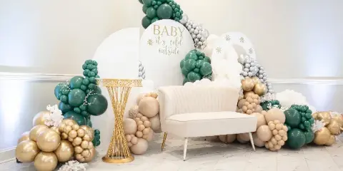 A professionally styled event backdrop featuring a green, gold, and silver balloon arch, a white velvet loveseat, and a gold pedestal table.