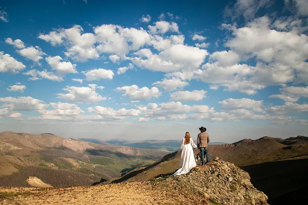 A bride and groom standing on a rocky mountain ridge overlooking a valley under a blue sky with white clouds.