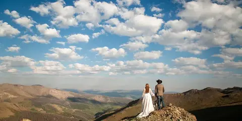 A bride and groom standing on a rocky mountain ridge overlooking a valley under a blue sky with white clouds.