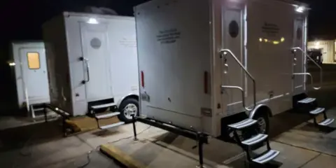 White portable restroom trailers with steps and exterior lighting parked at night.