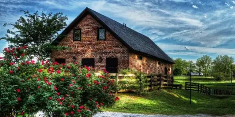 Exterior view of a historic red brick barn with a black metal roof, surrounded by green grass, a wooden fence, and blooming red rose bushes under a blue sky.