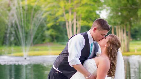 A bride and groom kissing in front of a pond with a fountain and trees.
