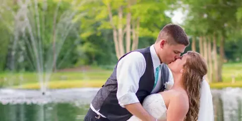 A bride and groom kissing in front of a pond with a fountain and trees.