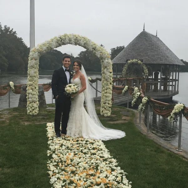 A bride and groom standing under a large floral arch with a flower-lined aisle at a waterfront venue.